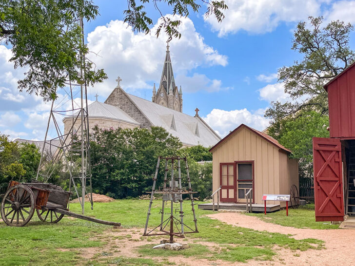 A red and white barn with an old carriage in front of them on a field while a cathedral can be seen behind surrounded by trees under bright blue skies with some white clouds, a picturesque scene on a Texas Hill Country road trip.