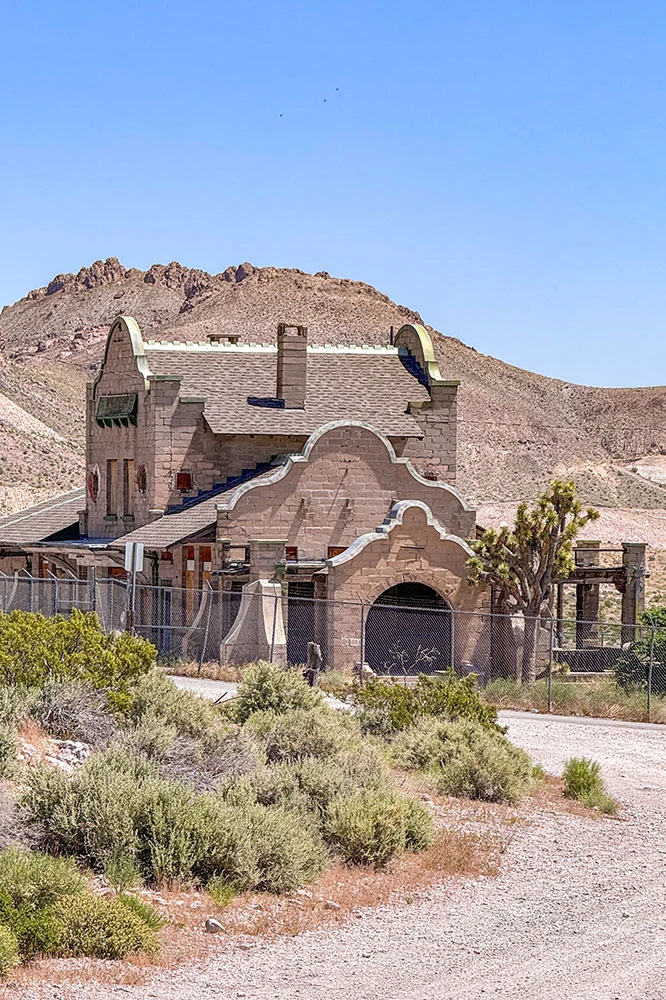 An old bricked house surrounded by bushes and mountain in the background under clear blue skies in Rhyoline Ghost Town, a destination in one of the road trips in nevada